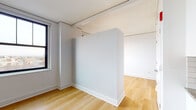 This single-floor unit presents a clean, contemporary design characterized by a consistent neutral palette and light hardwood flooring that runs throughout the living areas. The open layout is defined by crisp white walls and a structural half-wall partition that delineates separate functional zones while maintaining a sense of openness. Natural light enters through black-framed windows, offering views of the surrounding urban streetscape.

The kitchen is efficiently arranged in a galley configuration, featuring white cabinetry with matte black hardware, granite countertops, a subway tile backsplash, and a full suite of stainless steel appliances including a range, microwave, and dishwasher. A bold black entry door adds a striking contrast at the unit's entrance.

The bedroom areas are well-proportioned, each benefiting from individual windows that bring in natural light and views of the neighboring architecture. The rooms are finished with the same light hardwood flooring and white walls, creating a bright and adaptable environment.

The bathroom is finished with classic white subway tile surrounding a soaking tub and shower, complemented by large-format gray floor tiles, a wall-mounted vanity with chrome fixtures, and a generously sized mirror above the sink, completing the unit's modern, functional aesthetic.