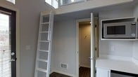 This thoughtfully designed space makes excellent use of vertical space through an elevated loft area, accessible via a white wooden ladder fixed along the wall. The soaring ceiling height creates an airy, open atmosphere despite the compact footprint, allowing the loft to function as an additional usable area above the main living space. The interior features neutral gray walls throughout, contributing to a clean, modern aesthetic. Dark hardwood-style flooring anchors the space and provides a warm contrast to the light-toned walls and white trim details. A doorway visible from the main area leads to a secondary space, likely a closet or utility area, illuminated by warm overhead lighting. A floor-level air vent indicates a central HVAC system for year-round climate control. Large windows positioned near the top of the loft level allow natural light to filter into the upper portion of the space, enhancing the sense of openness. The white ladder, door frames, and trim work are consistent throughout, lending a cohesive, well-finished look. Overall, the space is designed to maximize functionality within a compact layout through clever vertical design.