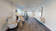 Located at 3420 Legacy Eagle Dr in Mount Pleasant, South Carolina, this single-floor community lounge offers a refined and welcoming environment defined by clean architectural lines and a coastal-contemporary aesthetic. The open, elongated layout is anchored by patterned carpet flooring that adds texture and warmth throughout the space.

Natural light pours in through multiple large windows, offering views of the surrounding community and creating a bright, airy atmosphere. The walls are finished in crisp white, providing a neutral backdrop that highlights the carefully curated décor, including large-scale abstract artwork, sculptural accents, and a striking open shelving unit displaying decorative objects with coastal-inspired color tones of teal and navy.

Seating areas feature modern wingback chairs and a metallic pedestal side table, arranged to encourage conversation and relaxation. Pendant and linear LED lighting fixtures hang from the ceiling, adding contemporary visual interest and supplementing the abundant natural light.

At one end of the space, a smooth stainless steel elevator and an open staircase with dark metal railings and wood treads provide access to upper levels of the building. The transition between light hardwood flooring near the entry and the patterned carpet in the lounge area subtly defines distinct zones within the space. Overall, the lounge presents a polished, welcoming atmosphere suited for community gathering and leisure.