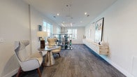 Located at 3420 Legacy Eagle Dr in Mount Pleasant, South Carolina, this single-floor community lounge offers a refined and welcoming environment defined by clean architectural lines and a coastal-contemporary aesthetic. The open, elongated layout is anchored by patterned carpet flooring that adds texture and warmth throughout the space.

Natural light pours in through multiple large windows, offering views of the surrounding community and creating a bright, airy atmosphere. The walls are finished in crisp white, providing a neutral backdrop that highlights the carefully curated décor, including large-scale abstract artwork, sculptural accents, and a striking open shelving unit displaying decorative objects with coastal-inspired color tones of teal and navy.

Seating areas feature modern wingback chairs and a metallic pedestal side table, arranged to encourage conversation and relaxation. Pendant and linear LED lighting fixtures hang from the ceiling, adding contemporary visual interest and supplementing the abundant natural light.

At one end of the space, a smooth stainless steel elevator and an open staircase with dark metal railings and wood treads provide access to upper levels of the building. The transition between light hardwood flooring near the entry and the patterned carpet in the lounge area subtly defines distinct zones within the space. Overall, the lounge presents a polished, welcoming atmosphere suited for community gathering and leisure.