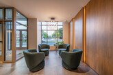 This single-floor lobby space showcases a polished, contemporary design characterized by large-format light grey porcelain tile flooring that extends throughout the entire area. The layout is thoughtfully organized into distinct functional zones, beginning with a bright entry vestibule framed by floor-to-ceiling glass doors and expansive windows that flood the space with natural light and offer views of the surrounding streetscape. A seating area near the entrance features sculptural teal lounge chairs arranged beneath a decorative pendant chandelier, creating a welcoming arrival experience.

Moving deeper into the lobby, a sleek reception desk with a dark countertop and warm wood base paneling is set against a striking textured stone accent wall with integrated LED strip lighting. Pendant lights in brass and tapered grey tones add visual interest above the desk. The elevator bank area transitions to a warmer palette, with rich wood-paneled walls, dark-framed elevator doors, and Edison-style exposed bulb ceiling fixtures. Framed artwork lines the corridor walls, adding a curated, gallery-like quality to the space. Throughout, the interplay of natural stone textures, warm wood tones, and cool grey finishes creates a refined, sophisticated ambiance suited to a high-end residential or commercial building.