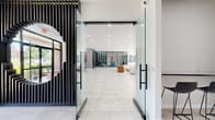 Located at 6806 Milbrook Park Dr in Baltimore, MD, this single-floor facility presents a thoughtfully designed, contemporary interior that balances open communal areas with dedicated functional spaces. Entry is made through full-height glass doors framed by striking black vertical slat panels, establishing a bold architectural tone from the outset. Large-format white porcelain tile flooring extends throughout, lending a clean, cohesive aesthetic to the entire space.

The expansive main lounge serves as the heart of the facility, furnished with white modular sofas and warm cognac accent chairs arranged beneath oversized circular pendant lights. Floor-to-ceiling glass sliding doors open directly to an outdoor green area, flooding the interior with natural light. An adjacent computer lounge features a wall-mounted wood-topped workstation with multiple desktop terminals, illuminated by a row of matte black pendant lights and accented with vibrant pop-art wall pieces.

A dedicated office area provides workstations and glass-partitioned private offices, offering a professional environment suited for administrative functions. The fitness center, enclosed in floor-to-ceiling glass, is well-equipped with ellipticals, treadmills, cycling equipment, free weights, and exercise balls, all beneath a warm wood slat ceiling with linear LED lighting. A secondary corridor with a bold vertical slat accent wall leads to additional private rooms, completing this versatile, modern facility.