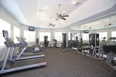 This fitness center offers a comprehensive workout environment spread across a single level, thoughtfully designed to accommodate a wide range of exercise needs. The main workout area features a tray ceiling with recessed lighting and ceiling fans, contributing to both visual appeal and air circulation during workouts. Neutral-toned walls and patterned carpet flooring provide a clean, functional aesthetic throughout the space.

The strength training section is well-appointed with a variety of cable machines, weight benches, and a dedicated dumbbell rack, complemented by large wall-mounted mirrors that visually expand the room. Multiple flat-screen TVs are positioned throughout, offering entertainment during workouts.

The cardio zone includes treadmills, ellipticals, and a recumbent bike, arranged near large windows fitted with blinds that allow natural light to filter in. Stability balls and additional accessories are available for stretching and supplemental exercises.

A water cooler station is conveniently placed within the facility, alongside posted fitness center rules. An adjacent room visible through a connecting doorway appears to offer a lounge or study area. An exterior entrance provides access to what appears to be an outdoor common area.