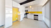 This single-story space showcases a distinctive mid-century modern aesthetic, defined by white tongue-and-groove ceilings, exposed dark structural beams, and a clean, minimal design sensibility throughout. The kitchen is a standout feature, pairing flat-front white cabinetry with a vibrant yellow backsplash and accent wall that adds a striking pop of color. Stainless steel appliances, including a range, over-the-range microwave, and dishwasher, complement the light-toned flooring, creating a functional and visually dynamic cooking space. A peninsula provides additional counter space and casual seating potential.

The bedrooms carry the architectural character of the unit, with the same white paneled ceilings and dark exposed beams continuing throughout. Each room features plush wall-to-wall carpeting, mirrored sliding closet doors, and neutral walls that contribute to a bright, airy atmosphere. Natural light enters through windows offering views of the surrounding neighborhood.

The bathroom maintains the space's clean, white palette with a tub-shower combination accented by white tile, a broad vanity counter with an integrated sink, chrome fixtures, and a large mirror. Overall, the space offers a cohesive, well-maintained interior with timeless mid-century character.