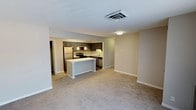 This single-floor residence showcases an open-concept design that connects the living area and kitchen into one cohesive, functional space. The living area is finished with neutral-toned carpeting that adds warmth and comfort underfoot, while light beige walls contribute to a bright, clean aesthetic throughout.

The kitchen is defined by dark cabinetry that provides a striking contrast against light countertops, creating a modern, polished look. A peninsula with a white base extends into the space, offering additional prep area and serving as a natural divider between the kitchen and living zones. Stainless steel appliances, including a refrigerator, range, and dishwasher, are seamlessly integrated into the cabinetry. Pendant lighting above the kitchen area adds a refined decorative touch, while recessed ceiling vents ensure efficient climate control throughout.

A doorway visible from the living area hints at additional rooms beyond, suggesting a well-organized floor plan. The overall ambiance is clean, contemporary, and move-in ready, with a neutral palette that offers flexibility for a variety of interior styles.