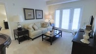 This single-floor residence offers a warm and inviting atmosphere defined by a clean, neutral color palette and thoughtfully curated design elements. The living area serves as the heart of the space, featuring plush carpeting underfoot and a comfortable cream-toned sofa accented with decorative pillows. A dark-finished coffee table anchors the seating arrangement, complemented by matching end tables and a coordinating media console that together create a cohesive and polished aesthetic. Soft ambient lighting from elegant table lamps enhances the room's welcoming ambiance. Two framed artworks are symmetrically displayed on the walls, adding a refined decorative touch. A standout feature of the living area is the set of French doors, which flood the space with generous natural light and provide direct access to the exterior, creating a seamless indoor-outdoor connection. The tray ceiling detail adds architectural interest and a sense of added height to the room. Overall, the space presents a harmonious blend of comfort and style, with every element contributing to a serene and livable environment suited for everyday relaxation and casual entertaining.