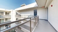 Located at 401 S La Fayette Park Place in Los Angeles, this rooftop sun deck is a generously sized open-air amenity space designed for communal outdoor living. The deck is framed by clean, modern architecture featuring crisp white stucco walls and sleek metal mesh railings that line the perimeter walkways, providing both safety and an industrial-contemporary aesthetic.

The space is organized into distinct functional zones. A partially shaded lounge area is outfitted with plush blue sofas and side tables, draped beneath a large shade sail that filters the California sun. String lights are strung overhead, creating an inviting ambiance for evening gatherings. An outdoor dining area features a dark table with coordinating chairs, ideal for al fresco meals.

A dedicated grilling station with a full-size gas barbecue makes this deck well-suited for entertaining. Additional dining tables and chairs are positioned nearby, offering ample seating for residents and guests. The deck also benefits from access to elevator and stairwell infrastructure, visible along the surrounding walkways that overlook a central courtyard below.

Unobstructed views of the Los Angeles cityscape extend from the deck's outer edges, offering a sweeping urban panorama beneath open skies. The polished concrete flooring and minimalist white architecture reflect the sunny Southern California setting throughout.