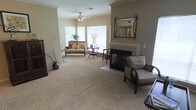 This single-floor living space features a generously sized living room with a warm, neutral color palette that creates an inviting and comfortable atmosphere. The room is anchored by a classic fireplace with a light-toned mantel, accented by a decorative wrought-iron mirror mounted above, adding an elegant focal point to the space. Plush wall-to-wall carpeting in a soft beige tone runs throughout, contributing to the room's cozy feel underfoot.

A ceiling fan with a light fixture is centrally positioned, providing both practical ventilation and ambient lighting. Multiple windows along one wall allow natural light to filter into the space, brightening the interior and offering views of surrounding greenery outside. The layout accommodates a comfortable seating arrangement, with ample room for both a loveseat and accent chairs, suggesting versatile furniture placement options.

Warm beige walls complement the carpeting and furnishings seamlessly. Decorative elements such as ornamental branches in a vase and a tripod floor lamp add character without overwhelming the space. Overall, the living room presents a relaxed yet refined aesthetic suited for everyday living.
