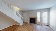 This two-story home in Saint Louis, Missouri, offers a well-organized layout that distributes living and private spaces across two distinct levels. The main floor centers around a comfortable living area finished with warm hardwood-style flooring and a textured ceiling. A tile-surround fireplace with a black firebox serves as a focal point, while a glass-paneled exterior door admits natural light and provides outdoor access. A white-railed staircase leads to the upper level, contributing an open, airy feel to the main living space. An additional open area on the main floor, also featuring hardwood-style flooring, connects to what appears to be a utility or storage space, adding functional versatility. The kitchen is outfitted with warm oak cabinetry, light countertops, a double stainless steel sink with a black faucet, and black appliances including a refrigerator, range, and microwave, all set atop tile flooring. Upstairs, carpeted rooms provide a quieter, more comfortable atmosphere. A ceiling fan with a light fixture is present in the upper-level space, which also opens to a hallway leading to a bathroom. The bathroom features an oak vanity with a white countertop, a large mirror, and tile flooring, maintaining consistent warm-toned finishes throughout the home.