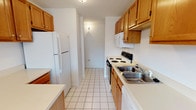 Located at 33 Pope Way in South Charleston, West Virginia, this single-floor residence offers a practical and well-organized layout. The galley-style kitchen is efficiently designed with warm oak cabinetry — including upper and lower storage — white laminate countertops, white tile flooring, and a full suite of appliances including a refrigerator, electric range, over-the-range microwave, and double-basin stainless steel sink. A white-painted brick accent wall adds texture as the kitchen transitions toward a connecting hallway. The open living and dining area features neutral carpet, a chandelier, and a window that brings in natural light, with the same white-painted brick wall providing a distinctive architectural detail. The bedrooms are carpeted and finished with clean white walls, each benefiting from natural light through windows with blinds and ceiling-mounted ventilation. The bathroom is clean and functional, with white tile flooring, a wood-toned vanity with countertop, a standard tub-shower combination, and crisp white fixtures throughout. The overall interior presents a straightforward, move-in-ready environment with consistent neutral tones and practical finishes across all areas.