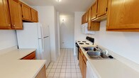 Located at 33 Pope Way in South Charleston, West Virginia, this single-floor residence offers a practical and well-organized layout. The galley-style kitchen is efficiently designed with warm oak cabinetry — including upper and lower storage — white laminate countertops, white tile flooring, and a full suite of appliances including a refrigerator, electric range, over-the-range microwave, and double-basin stainless steel sink. A white-painted brick accent wall adds texture as the kitchen transitions toward a connecting hallway. The open living and dining area features neutral carpet, a chandelier, and a window that brings in natural light, with the same white-painted brick wall providing a distinctive architectural detail. The bedrooms are carpeted and finished with clean white walls, each benefiting from natural light through windows with blinds and ceiling-mounted ventilation. The bathroom is clean and functional, with white tile flooring, a wood-toned vanity with countertop, a standard tub-shower combination, and crisp white fixtures throughout. The overall interior presents a straightforward, move-in-ready environment with consistent neutral tones and practical finishes across all areas.