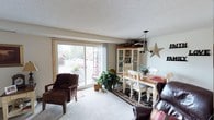 This single-floor home offers a warm and inviting layout designed for comfortable everyday living. The combined living and dining area serves as the heart of the home, featuring plush carpeting, a textured ceiling, and a large sliding glass door that floods the space with natural light and provides views of the outdoors. The dining area is anchored by a warm wood table surrounded by white chairs, a classic chandelier overhead, and a white china cabinet displaying decorative dishware — all complemented by charming wall décor that adds a personal, homey touch.

The kitchen is compact yet well-equipped, featuring rich dark wood cabinetry, light countertops, tile backsplash, and essential appliances including an electric range, refrigerator, and dishwasher. A window above the sink brings in additional natural light.

The home includes three bedrooms, each carpeted and finished with textured ceilings. Two of the bedrooms feature windows dressed with curtains that provide both privacy and soft light, while the third offers a cozier, more intimate feel. Each room provides comfortable accommodations with ample space for furnishings.

A full bathroom with tile flooring, a vanity with wood cabinetry, and a tub-shower combination completes the layout. Throughout the home, a consistent neutral color palette and warm accents create a cohesive, lived-in atmosphere.