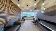 Located at 2255 Rue St-Mathieu in Montréal, this refined lounge space offers a carefully curated blend of modern design and warm, inviting materials. The interior is anchored by a striking full-height slatted wood accent wall that introduces natural warmth and texture, contrasting elegantly with the charcoal-toned walls adorned with an abstract mural-style finish.

The main seating area features a leather sofa and a pair of sleek leather lounge chairs arranged around small side tables, creating an intimate social setting. A contemporary linear fireplace is integrated into the feature wall, adding both ambiance and a focal point to the room. Globe pendant lights suspended from the ceiling provide a warm, layered glow throughout the space.

Large floor-to-ceiling windows and a glazed balcony surround allow abundant natural light to flood the interior while framing sweeping panoramic views of the Montréal skyline — a standout architectural feature of the lounge. The light stone-tiled flooring runs throughout, lending a clean, cohesive aesthetic underfoot.

A functional kitchenette area is discreetly integrated near the entrance, complete with gray cabinetry, a white quartz countertop, and a sink — offering convenience without disrupting the lounge's refined atmosphere. The space is completed by a dedicated entry zone with controlled glass door access, ensuring a polished and well-appointed experience from arrival to departure.