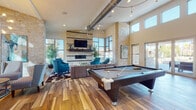 This expansive single-floor communal lounge is designed to accommodate a variety of social and recreational activities within one open, flowing space. Soaring ceilings with exposed industrial-style ductwork lend a modern, loft-inspired character, while large multi-pane windows flood the interior with abundant natural light and offer views of the surrounding landscape.

Anchoring one end of the space is a striking floor-to-ceiling light brick fireplace wall, complete with a wall-mounted flat-screen television and floating shelves. Vivid teal accent chairs and a cowhide-patterned area rug define the lounge seating area, creating an inviting focal point. Warm, wide-plank hardwood-style flooring runs throughout, unifying each zone cohesively.

A full-size billiards table occupies a central position in the room, offering a key recreational feature. Nearby, a high-top dining table with bar stools provides a casual gathering spot. Adjacent to the entrance, a dedicated workstation area features a long granite-topped counter lined with seating, backed by a bold, colorful mural in teal, yellow, and orange. A kitchenette with dark cabinetry and appliances completes the functional amenities of this well-appointed space.