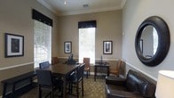 This single-floor space functions as a combined business center and lounge area, designed to offer both productivity and comfort. The room is finished with warm neutral-toned walls, white crown molding, and a white chair rail that adds a classic architectural touch throughout.

Natural light flows generously into the space through multiple windows dressed with white horizontal blinds and dark valances accented with decorative hardware, creating a bright yet polished atmosphere. Framed artwork on the walls adds a refined, welcoming character to the room.

The business center area features dedicated computer workstations set on sleek dark desks, accompanied by tan upholstered chairs, providing a functional workspace for focused tasks. A large dark wood conference-style table with a full set of matching chairs serves as the centerpiece of the room, ideal for collaborative work or group meetings.

A comfortable dark leather sofa is positioned in the lounge area, offering a more relaxed seating option. The flooring features a neutral patterned carpet that ties the space together cohesively. Recessed lighting complements the natural light, ensuring the room remains well-lit throughout the day. Overall, the space strikes a balance between a professional and comfortable environment.