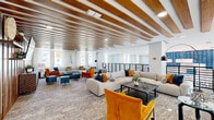 This sophisticated two-floor communal space offers a generous and thoughtfully designed environment across a spacious main level and a more intimate upper mezzanine. The architectural centerpiece is an eye-catching ceiling treatment of warm wood slat beams set against crisp white surfaces, creating a rhythm of texture and warmth that carries throughout both floors. The expansive main level is arranged as an open lounge, furnished with large sectional sofas in neutral tones, complemented by bold accent chairs in rich amber, teal, and orange velvet upholstery. Multiple seating vignettes are anchored by area rugs and low coffee tables, making the space ideal for both relaxed gatherings and social interaction. A large wall-mounted display screen and a distinctive textured wood media console add a media-lounge element to one end of the room. Abundant natural light pours in through tall windows dressed in sheer white curtains, while dramatic arc floor lamps with matte black shades punctuate the corners. The upper mezzanine level, connected by a sleek staircase with black metal railings, overlooks the main floor through an open balustrade, revealing double-height ceilings and a vibrant blue tile accent wall below. The lower level features additional seating, greenery, and display areas, contributing to an energetic yet welcoming ambiance throughout.
