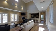 This single-floor clubhouse is designed to foster community gathering in a warm and stylish environment. The main lounge area immediately impresses with its dramatic vaulted ceiling that rises to a striking height, amplifying the sense of openness throughout the space. Natural light pours in through tall windows and glass-paned French doors, illuminating the light wood flooring that runs throughout.

A floor-to-ceiling stacked stone fireplace serves as the room's focal centerpiece, framed by a wooden mantel and topped with a wall-mounted television — creating an inviting spot for relaxation. The seating area is thoughtfully arranged with a plush sofa, accent chairs, and a rustic wooden coffee table set atop a patterned area rug.

Adjacent to the lounge, an open kitchen features dark cabinetry, stainless steel appliances, and a countertop bar with stools — ideal for casual socializing. A separate dining area accommodates larger groups with a generous wooden table and bench seating, providing a more formal setting for community meals or events.

Wall-mounted sconce lighting adds a warm, ambient glow, while recessed ceiling lights ensure the space remains bright and functional. The overall ambiance blends rustic charm with modern comfort, making this clubhouse a versatile and inviting destination for residents.