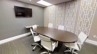 This professional conference room is thoughtfully designed to facilitate productive meetings in a polished and comfortable environment. At the center of the room sits a large, dark espresso-finish oval conference table surrounded by white ergonomic chairs with sleek chrome bases, offering seating for up to eight attendees. A wall-mounted flat-screen display is positioned prominently on the soft grey wall, ensuring clear visibility from all seating positions and supporting seamless presentations and video conferencing. One wall features a striking geometric-patterned wallcovering in warm taupe and white tones, lending the room a refined, contemporary aesthetic that balances professionalism with visual interest. A glass-legged side desk or credenza is tucked neatly against the wall, providing additional workspace or surface for equipment. The flooring consists of grey-toned textured carpet, adding warmth and acoustic comfort to the space. Overhead, a flush skylight-style ceiling panel provides bright, even illumination throughout the room. The overall ambiance is clean, modern, and professional — well-suited for client meetings, team collaborations, and presentations alike.