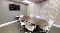 This professional conference room is thoughtfully designed to facilitate productive meetings in a polished and comfortable environment. At the center of the room sits a large, dark espresso-finish oval conference table surrounded by white ergonomic chairs with sleek chrome bases, offering seating for up to eight attendees. A wall-mounted flat-screen display is positioned prominently on the soft grey wall, ensuring clear visibility from all seating positions and supporting seamless presentations and video conferencing. One wall features a striking geometric-patterned wallcovering in warm taupe and white tones, lending the room a refined, contemporary aesthetic that balances professionalism with visual interest. A glass-legged side desk or credenza is tucked neatly against the wall, providing additional workspace or surface for equipment. The flooring consists of grey-toned textured carpet, adding warmth and acoustic comfort to the space. Overhead, a flush skylight-style ceiling panel provides bright, even illumination throughout the room. The overall ambiance is clean, modern, and professional — well-suited for client meetings, team collaborations, and presentations alike.