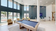 This contemporary residential clubhouse is designed to impress from the moment you enter. The grand atrium lobby sets the tone with its soaring double-height ceilings, a striking indoor tree planter encased in mosaic tile, and an open floating staircase with sleek metal railings that leads to an upper mezzanine level overlooking the main floor. Large-format light tile flooring runs throughout, lending a clean, cohesive aesthetic to the entire space.

The expansive main lounge area features a bold central fireplace, a generous curved sectional sofa, and ample seating arranged for both socializing and relaxation. Tall columns and recessed lighting complement the open, airy layout, while an upper-level railing adds architectural depth. A dedicated game lounge showcases a full-size pool table set against deep teal accent walls, with floor-to-ceiling windows offering views of the resort-style outdoor pool and surrounding community.

Additional gathering spaces include a casual dining area with a woven pendant light and teal accent wall, a bright lounge with large windows and curated art, a media nook with a wall-mounted TV, and a well-lit reception corridor. Throughout, the design balances modern minimalism with warm, inviting touches — creating a welcoming and functional community hub.