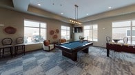 This single-floor community amenity space is thoughtfully designed to encourage socializing and recreation. The main game room is generously sized, featuring a full-size billiards table positioned beneath an industrial-style pendant light fixture, and a regulation shuffleboard table set against a textured houndstooth-patterned accent wall with a wall-mounted flat-screen TV above. Patterned carpet tiles in muted blue and gray tones cover the game area floor, adding a modern, layered aesthetic throughout. Large windows on multiple walls flood the space with natural light and provide views of the surrounding exterior. Comfortable accent seating, including upholstered chairs with warm-toned pillows, is arranged around the room for relaxed conversation. Adjacent to the game area, the kitchenette and bar area transitions to rich hardwood flooring and features a striking decorative tile backsplash, stainless steel refrigerator, white cabinetry with open wood shelving, a dark countertop bar, and elegant globe pendant lights overhead. Stylish round-back barstools line the bar counter, creating a casual yet refined gathering spot. Decorative wood slice wall art adds a natural, organic touch to the warm neutral-toned walls, tying the overall design together with an inviting and contemporary feel.