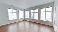 This single-floor space is defined by its clean, minimalist design and an abundance of natural light. The main living area features warm hardwood flooring that contrasts elegantly with the crisp white walls and bright white trim throughout. A standout feature of the space is the wraparound arrangement of large, multi-pane windows that line two walls, flooding the interior with daylight and offering sweeping views of the surrounding tree line. The windows are fitted with simple roller blinds, providing privacy while maintaining the room's airy, open atmosphere. The neutral gray-white palette of the walls creates a versatile backdrop suitable for a variety of design styles. Ceiling-mounted HVAC vents indicate a modern climate control system, adding to the functional appeal of the space. The open layout provides generous room to configure furniture and décor to suit multiple uses. The combination of natural light, hardwood floors, and expansive windows gives the space a welcoming, serene ambiance throughout.