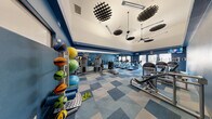 Located at 9550 W Sahara Ave in Las Vegas, Nevada, this fitness center offers a well-equipped exercise space designed to support a comprehensive workout experience. The room features soaring vaulted ceilings that lend an open, airy feel, accented by striking geometric ceiling fixtures and ceiling fans that keep the environment comfortable during intense sessions.

The interior is styled with bold, deep blue walls paired with a patterned blue and grey modular carpet flooring, creating a dynamic and energetic atmosphere. Large windows and a glass door along one wall allow natural light to fill the space and provide views of the surrounding exterior.

Full-length mirrors line one wall, visually expanding the room and allowing users to monitor their form during exercise. The cardio section is well-stocked with multiple treadmills and elliptical machines arranged in rows for ample personal space. A dedicated strength training area features a cable and weight machine station, along with a rack of medicine balls and stability balls in various sizes, catering to functional fitness routines. Additional strength machines are positioned throughout for targeted muscle training.

A mounted television and clock are visible near the entrance, contributing to a practical and user-friendly environment. The fitness center is thoughtfully organized to maximize utility across its footprint, making it a capable and inviting facility for residents or members seeking a well-rounded exercise space.
