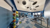 Located at 9550 W Sahara Ave in Las Vegas, Nevada, this fitness center offers a well-equipped exercise space designed to support a comprehensive workout experience. The room features soaring vaulted ceilings that lend an open, airy feel, accented by striking geometric ceiling fixtures and ceiling fans that keep the environment comfortable during intense sessions.

The interior is styled with bold, deep blue walls paired with a patterned blue and grey modular carpet flooring, creating a dynamic and energetic atmosphere. Large windows and a glass door along one wall allow natural light to fill the space and provide views of the surrounding exterior.

Full-length mirrors line one wall, visually expanding the room and allowing users to monitor their form during exercise. The cardio section is well-stocked with multiple treadmills and elliptical machines arranged in rows for ample personal space. A dedicated strength training area features a cable and weight machine station, along with a rack of medicine balls and stability balls in various sizes, catering to functional fitness routines. Additional strength machines are positioned throughout for targeted muscle training.

A mounted television and clock are visible near the entrance, contributing to a practical and user-friendly environment. The fitness center is thoughtfully organized to maximize utility across its footprint, making it a capable and inviting facility for residents or members seeking a well-rounded exercise space.