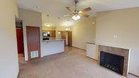 This single-floor space offers an open and airy layout highlighted by soaring vaulted ceilings that lend a sense of generous volume to the living area. A wood-toned ceiling fan with an integrated light fixture anchors the main living space, complementing the warm, neutral tones of the carpeted floors. A tiled fireplace with a classic surround serves as a focal point on one side of the room, adding a cozy and inviting ambiance.

The open-concept design flows seamlessly into the kitchen, which features warm wood cabinetry, a stainless steel refrigerator, and a built-in microwave above the range. A raised breakfast bar with a stone or granite-style countertop provides a natural division between the kitchen and living area while maintaining the open feel. Pendant lighting hangs above the bar and kitchen, offering both functional illumination and a decorative touch that enhances the warmth of the space.

Tile flooring in the kitchen and entry areas contrasts tastefully with the carpeted living space. The layout is practical and well-connected, with clear sightlines throughout. Overall, the space presents a comfortable and functional environment with architectural details that elevate its everyday livability.
