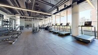 Located at 74 Bathurst Street in Toronto's vibrant King West neighbourhood, this fitness facility spans a generously sized single floor, offering a comprehensive and well-equipped workout environment. The space is defined by an industrial-inspired aesthetic, featuring an exposed ceiling with dark-painted ductwork, structural columns, and sleek linear lighting that runs the length of the floor, creating a bright and energetic atmosphere.

Floor-to-ceiling windows line one entire side of the facility, flooding the space with abundant natural light and providing sweeping views of the Toronto skyline — an especially motivating backdrop for cardio workouts. A full row of treadmills, ellipticals, stair climbers, and stationary bikes is positioned directly along this glazed perimeter to maximize the view.

The strength training zone is well-appointed with multiple cable machines, squat racks, a comprehensive dumbbell rack, and adjustable benches, all set on durable rubber flooring. A dedicated studio area enclosed by full-height glass walls offers a versatile space for group classes or focused training, complete with stability balls and a large display screen. Wide mirrors throughout the main floor further enhance the sense of openness. The facility also includes change room access, making it a fully self-contained fitness destination.