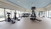 This fitness center offers a spacious, thoughtfully designed workout environment that caters to a wide range of exercise needs. The facility features a clean, contemporary aesthetic with a light blue ceiling and neutral-toned walls that create a calm, energizing atmosphere. Large floor-to-ceiling windows line the perimeter, flooding the space with abundant natural light and providing open views of the surrounding greenery, making workouts feel less confined and more invigorating. The flooring consists of durable, textured rubber matting, providing both safety and comfort underfoot. Cardio equipment is prominently featured, with a row of treadmills, stationary bikes, and elliptical machines positioned to take advantage of the natural light. The strength training area is well-stocked with a Motion multi-use cable and functional trainer station, a full rack of kettlebells and medicine balls of varying weights, and additional free weights. Full-length mirrors are mounted along one wall, enhancing the sense of space and allowing users to monitor their form. Recessed overhead lighting supplements the natural light, ensuring the facility is well-lit at all hours. The open floor plan allows for easy movement between equipment zones, making this fitness center a comprehensive and welcoming space for all fitness levels.
