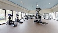 This fitness center offers a spacious, thoughtfully designed workout environment that caters to a wide range of exercise needs. The facility features a clean, contemporary aesthetic with a light blue ceiling and neutral-toned walls that create a calm, energizing atmosphere. Large floor-to-ceiling windows line the perimeter, flooding the space with abundant natural light and providing open views of the surrounding greenery, making workouts feel less confined and more invigorating. The flooring consists of durable, textured rubber matting, providing both safety and comfort underfoot. Cardio equipment is prominently featured, with a row of treadmills, stationary bikes, and elliptical machines positioned to take advantage of the natural light. The strength training area is well-stocked with a Motion multi-use cable and functional trainer station, a full rack of kettlebells and medicine balls of varying weights, and additional free weights. Full-length mirrors are mounted along one wall, enhancing the sense of space and allowing users to monitor their form. Recessed overhead lighting supplements the natural light, ensuring the facility is well-lit at all hours. The open floor plan allows for easy movement between equipment zones, making this fitness center a comprehensive and welcoming space for all fitness levels.