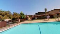 This outdoor amenity area centers around a generously sized swimming pool with clear turquoise water, set within a residential complex. The pool is enclosed by a dark wrought-iron safety fence, providing both security and visibility into the well-maintained aquatic space. A concrete pool deck surrounds the perimeter, offering space for poolside seating, with at least one bench visible along the edge for relaxation.

Safety equipment, including a life ring, is mounted near the pool area, reflecting a practical and safety-conscious design. The surrounding landscape includes mature trees that provide natural shade and greenery, contributing to a pleasant outdoor atmosphere.

The residential buildings framing the pool are two-story structures with earthy brown and tan exteriors, featuring exterior stairways and covered walkways. The architectural style is consistent with a mid-century multi-unit residential design. The complex benefits from an abundance of natural sunlight, with clear blue skies enhancing the open and airy feel of the outdoor space. Overall, the pool area serves as a central communal feature of the property, offering a refreshing retreat within the complex.