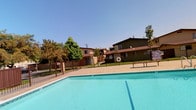 This outdoor amenity area centers around a generously sized swimming pool with clear turquoise water, set within a residential complex. The pool is enclosed by a dark wrought-iron safety fence, providing both security and visibility into the well-maintained aquatic space. A concrete pool deck surrounds the perimeter, offering space for poolside seating, with at least one bench visible along the edge for relaxation.

Safety equipment, including a life ring, is mounted near the pool area, reflecting a practical and safety-conscious design. The surrounding landscape includes mature trees that provide natural shade and greenery, contributing to a pleasant outdoor atmosphere.

The residential buildings framing the pool are two-story structures with earthy brown and tan exteriors, featuring exterior stairways and covered walkways. The architectural style is consistent with a mid-century multi-unit residential design. The complex benefits from an abundance of natural sunlight, with clear blue skies enhancing the open and airy feel of the outdoor space. Overall, the pool area serves as a central communal feature of the property, offering a refreshing retreat within the complex.