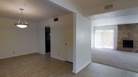 This single-story space presents a straightforward, functional layout with a clear division between zones. The entry area features light-toned hardwood-style flooring that extends through the central corridor, providing a clean and easy-to-maintain surface. A structural half-wall with an arched opening separates this area from the adjacent carpeted living space, which benefits from generous natural light streaming through a large sliding glass door or picture window along the far wall. The bright, neutral-toned carpeting in the living area complements the white walls throughout. Ceiling vents are visibly integrated into the layout, indicating a central HVAC system. The white walls are consistent throughout, contributing to a bright and neutral interior. A door in the corridor leads to a darker hallway or room beyond, suggesting additional private spaces within the unit. The textured ceiling is visible in portions of the space. Overall, the layout is practical and open, with a clear distinction between the hard-floored transitional areas and the softer, carpeted living zone, making efficient use of the available footprint.