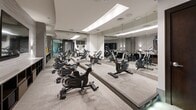 Located at 295 E Speer Blvd in Denver, Colorado, this dedicated cycling studio offers a polished, purpose-built environment for indoor cycling workouts. The room is outfitted with a full row of Matrix stationary spin bikes arranged to maximize use of the available floor space, providing an efficient and focused exercise setting.

The interior design is notably modern and refined. Full-length mirrors line one wall, visually expanding the space and allowing cyclists to monitor their form during workouts. Recessed linear lighting in the ceiling casts a clean, even illumination throughout the room, complementing the neutral gray tones of the textured wall panels and low-pile carpeted flooring.

Entry to the studio is through a glass door framed by dark wood paneling accented with a decorative mosaic tile wall feature, lending a boutique-gym aesthetic to the space. Along one wall, built-in open shelving provides storage, and a wall-mounted flat-screen television serves as a focal point for guided workouts or entertainment. Decorative accents in gold add subtle visual interest against the otherwise understated palette.

Ceiling-mounted speakers and ventilation fixtures ensure a comfortable and acoustically suitable environment. The overall ambiance is contemporary, clean, and purpose-driven—designed to support a premium indoor cycling experience in the heart of Denver.