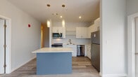 Located at 1 Royal Dr. in Somersworth, New Hampshire, this single-floor residence offers a thoughtfully designed layout with modern finishes throughout. The kitchen serves as a central highlight, featuring crisp white cabinetry extending to the ceiling, light quartz countertops, a slate-blue island with pendant lighting, and a full suite of stainless steel appliances including a refrigerator, range, microwave, and dishwasher. Light wood-style flooring ties the kitchen and adjacent living areas together seamlessly, and a glass door provides convenient access to the outdoors. The bedrooms are carpeted for comfort and bathed in natural light through large windows with views of the surrounding neighborhood and greenery. One bedroom includes a spacious walk-in closet outfitted with wire shelving systems for ample storage. The bathrooms are clean and contemporary — one features a walk-in shower with a white surround and wood-toned vanity with quartz countertops, while a second bathroom offers a similarly styled vanity with light flooring. Throughout the residence, neutral gray walls, white trim, and recessed lighting contribute to a bright, cohesive aesthetic that is both modern and welcoming.
