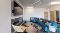 Located at 3124 W Broad St in Richmond, Virginia, this single-floor space combines functional design with modern aesthetics across several distinct areas. The open layout connects a comfortable lounge, a dedicated conference room, and a sleek kitchenette, creating a versatile environment suited for both collaboration and relaxation.

The lounge area serves as a central gathering space, anchored by a large teal sectional sofa adorned with colorful accent pillows. A wall-mounted flat-screen television sits above a linear tile-faced fireplace, providing a focal point for the room. Polished concrete floors run throughout, complemented by bold patterned area rugs that define each zone. A console table and round side tables complete the seating arrangement, while a yellow accent chair adds a pop of color.

The conference room offers a professional setting with a dark rectangular table accommodating up to eight, surrounded by a mix of charcoal and yellow upholstered chairs. Floor-to-ceiling windows flood the space with natural light and provide views of the surrounding streetscape.

The kitchenette features clean white cabinetry, quartz countertops, stainless steel appliances including a full-size refrigerator and under-counter unit, and a single-basin sink — providing practical support for events or daily use.