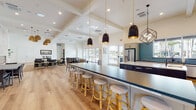 This single-floor community clubhouse is thoughtfully designed to offer a versatile, upscale gathering space with distinct functional zones flowing seamlessly across light-toned hardwood flooring. The open-plan layout benefits from soaring ceilings with coffered and beamed detailing, recessed lighting, and an abundance of natural light streaming through large floor-to-ceiling windows and glass doors that frame views of the outdoor pool and surrounding landscaping.

The fully equipped gourmet kitchen is a standout feature, showcasing a professional-grade stainless steel range with a sleek overhead hood, vibrant teal glass tile backsplash, gray shaker cabinetry, and dark countertops. A large central island with bar-height seating accommodates casual dining, while a full-size stainless steel refrigerator and ample counter space make the kitchen ideal for entertaining. Stylish pendant lighting in mixed black and geometric finishes adds a modern touch overhead.

Adjacent dining and lounge areas offer flexible seating arrangements, including upholstered chairs, woven area rugs, and multiple wall-mounted displays. A striking floor-to-ceiling stone fireplace anchors one end of the lounge, providing a warm focal point. The overall ambiance is bright, contemporary, and welcoming.