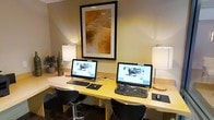 This business center offers a functional and focused environment designed to keep guests connected and productive. The space is anchored by a long, light-wood workstation desk that accommodates two full computer setups, each featuring an all-in-one desktop monitor, keyboard, and mouse — providing ample room for side-by-side use without feeling cramped. Ergonomic bar-height chairs in a sleek dark finish accompany each station, offering a comfortable seating arrangement for extended work sessions. Soft, warm lighting is provided by two table lamps positioned at either end of the desk, casting an inviting glow that complements the neutral, beige-toned walls. A framed piece of abstract artwork in warm amber and earth tones hangs prominently above the workstation, adding a touch of character and visual interest to the space. A small decorative plant and ceramic vase accent one corner of the desk, contributing to the room's polished yet welcoming atmosphere. The overall design balances practicality with comfort, making this business center a well-appointed spot for handling correspondence, printing, or any work-related tasks during a stay.