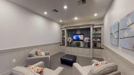 This theater room is thoughtfully designed to deliver a comfortable and stylish entertainment experience. The space is defined by its elegant built-in entertainment wall, which serves as the room's centerpiece. Finished in a soft gray tone, the unit features a centrally mounted flat-screen television framed by blue drape panels, flanked by open display shelving and a dark granite countertop with cabinetry below—combining functionality with refined aesthetics.

The room's walls are adorned with classic wainscoting, adding architectural depth and a polished, traditional character throughout. Soft gray carpet covers the floor, contributing to the room's acoustic comfort and inviting atmosphere. Recessed lighting in the ceiling provides even, ambient illumination across the space.

Seating consists of multiple plush gray upholstered armchairs accented with orange and white patterned throw pillows, arranged to face the entertainment wall. A tufted black leather ottoman anchors the seating area. Soft blue floral artwork decorates the side wall, adding a subtle touch of color.

A doorway connects the theater to an adjacent space with herringbone brick flooring and natural light, providing a seamless transition between the entertainment room and the rest of the home.