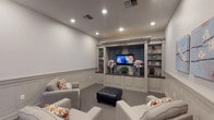 This theater room is thoughtfully designed to deliver a comfortable and stylish entertainment experience. The space is defined by its elegant built-in entertainment wall, which serves as the room's centerpiece. Finished in a soft gray tone, the unit features a centrally mounted flat-screen television framed by blue drape panels, flanked by open display shelving and a dark granite countertop with cabinetry below—combining functionality with refined aesthetics.

The room's walls are adorned with classic wainscoting, adding architectural depth and a polished, traditional character throughout. Soft gray carpet covers the floor, contributing to the room's acoustic comfort and inviting atmosphere. Recessed lighting in the ceiling provides even, ambient illumination across the space.

Seating consists of multiple plush gray upholstered armchairs accented with orange and white patterned throw pillows, arranged to face the entertainment wall. A tufted black leather ottoman anchors the seating area. Soft blue floral artwork decorates the side wall, adding a subtle touch of color.

A doorway connects the theater to an adjacent space with herringbone brick flooring and natural light, providing a seamless transition between the entertainment room and the rest of the home.