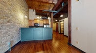 Located at 228–298 N Plankinton Ave in downtown Milwaukee, this single-floor loft-style space showcases a compelling blend of historic industrial architecture and modern finishes. The open-concept living area is defined by dramatic exposed timber beam ceilings with visible ductwork, warm hardwood floors, and expansive original brick walls in cream and mixed tones, all contributing to a rich, urban aesthetic.

The kitchen serves as a focal point of the main living space, featuring a striking teal beadboard peninsula with dark granite countertops, warm wood cabinetry, stainless steel appliances, and pendant lighting. The kitchen flows seamlessly into the generous living area, which benefits from natural light streaming through windows with city views and a ceiling fan overhead.

The bedroom continues the industrial character with soaring ceilings, prominent exposed brick walls, and carpeted flooring for comfort. A walk-in closet provides practical storage. Two bathrooms are present — one featuring an exposed brick accent wall, a vanity with dark countertops, and a tub-shower combination; the other offering a clean, tiled tub-shower setup. The overall ambiance is warm, character-rich, and distinctly urban.