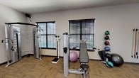Located at 5960 Siegen Ln in Baton Rouge, Louisiana, this single-floor fitness center is a well-appointed facility designed to support a comprehensive workout experience. The space is characterized by soaring ceilings, recessed lighting, and a bold, colorful geometric mural accent wall that adds a vibrant, energetic atmosphere to the room. Large windows and glass doors allow ample natural light to flow throughout, offering views of the surrounding outdoor landscape.

The cardio zone features a row of treadmills, elliptical machines, and a rowing machine, all positioned in front of wall-mounted flat-screen televisions for entertainment during workouts. An overhead ceiling fan provides additional air circulation across this area. A dedicated stretching and functional training zone includes interlocking floor mats, a weight bench, and ample open space.

The strength training section is outfitted with professional-grade Cybex cable and resistance machines, a full rack of dumbbells ranging across multiple weights, medicine balls on a wall-mounted rack, and stability balls. A large floor-to-ceiling mirror enhances the sense of space and allows users to monitor their form. The flooring throughout is durable, textured carpet tile suited for high-traffic fitness use. The facility is thoughtfully laid out to accommodate multiple users simultaneously across distinct training zones.