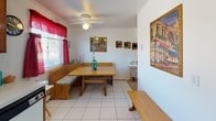 This single-floor home offers a practical and well-utilized layout across several distinct areas. The kitchen features warm wood cabinetry, white tile countertops with a tiled backsplash, and a window dressed with a red valance that allows natural light to brighten the workspace. Appliances and ample storage make the kitchen fully functional.

Adjacent to the kitchen, the dining area centers around a wood booth-style banquette with built-in bench seating and a matching table, creating a cozy nook ideal for casual meals. A ceiling fan with a light fixture overhead provides air circulation, while windows with red curtains bring in additional natural light. White tile flooring extends throughout this area.

The living room features neutral carpet, white walls, and a wood entertainment center. Two sofas provide comfortable seating, and wall décor adds a personalized touch. A wood entry door with a brass knob is visible from this space.

The bedroom is carpeted and equipped with a ceiling fan, natural window light, and ample room for a full bedroom suite including a dresser and accent chair. The bathroom completes the home with white tile flooring, a pedestal sink, toilet, and a window with a teal curtain accent.
