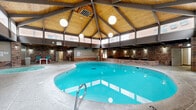 Located at 815 N 39th St in Grand Forks, North Dakota, this indoor pool and recreation area offers a well-appointed, single-floor facility designed for leisure and activity. The architectural character of the space is defined by striking vaulted wood-plank ceilings with exposed dark steel beams, warm brick walls, and clerestory windows that allow abundant natural light to flood the interior, creating an open and inviting atmosphere.

At the heart of the facility is a generously sized indoor swimming pool with a curved, freeform shape and gradual entry steps, surrounded by a textured concrete pool deck. Adjacent to the pool is a separate hot tub spa tucked into a more intimate brick-enclosed alcove, accompanied by high-top seating for relaxation.

The recreation area extends beyond the pool to include a carpeted zone featuring a full-size ping-pong table, ideal for active recreation. Multiple seating arrangements with tables and colorful chairs are distributed throughout the space, providing areas for socializing and lounging. A vending machine offers convenient refreshment access. Globe pendant light fixtures and wall-mounted sconces complement the natural light, lending warmth to the evenings.

Utility doors and clearly marked exits ensure practical circulation throughout the facility. The combination of recreational amenities, distinctive architectural details, and comfortable gathering spaces makes this a versatile and functional indoor recreation destination.