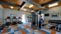 Located in Grapevine, TX, this single-floor fitness center is thoughtfully designed to inspire an active lifestyle. The space immediately draws attention with its striking coffered ceiling, finished in a warm amber-orange tone and framed by bold gray structural beams — a design choice that adds both architectural character and a sense of energy to the room. The flooring continues the vibrant color story, featuring a dynamic patchwork pattern of orange, blue, and gray tiles that give the space a lively, motivating atmosphere. The fitness area is well-equipped with a range of cardio machines, including multiple treadmills and an elliptical trainer, as well as a multi-station cable strength training unit to support a comprehensive workout routine. A wall-mounted flat-screen television provides entertainment during exercise sessions. Natural light filters in through windows on multiple walls, keeping the space bright and welcoming, while recessed and overhead lighting ensures consistent illumination throughout. Framed artwork adorns the walls, adding a polished, curated feel to the environment. The layout is open and spacious, allowing for comfortable movement between equipment. Overall, this fitness center combines functional amenities with an engaging visual design to create an uplifting workout environment.