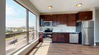 This single-floor unit is thoughtfully designed with a clean, contemporary aesthetic throughout. The kitchen serves as a central highlight, showcasing rich dark-wood cabinetry extending floor to ceiling, light-colored countertops, and a full suite of stainless steel appliances including an electric range, built-in microwave, dishwasher, and refrigerator. A double-basin sink sits beneath the cabinetry, and the layout provides generous counter space for meal preparation. Large floor-to-ceiling windows in the kitchen and adjacent living area flood the space with natural light and offer elevated views of the surrounding neighborhood and streetscape. Hardwood-style flooring runs throughout the main living and kitchen areas, contributing warmth and continuity to the space. The bedroom is carpeted for comfort and features multiple windows equipped with roller shades, along with a wall-mounted HVAC unit for climate control. Views of tree-lined streets and neighboring structures are visible from the bedroom windows. The bathroom is finished with a dark-wood vanity, white countertop, a full-size mirror, and a tub-shower combination. Neutral gray wall tones are consistent throughout, creating a cohesive and understated modern ambiance across the entire unit.
