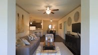 This single-floor residence showcases an open-concept design that connects the living area to the kitchen, creating a spacious and cohesive layout. The living room serves as the centerpiece, featuring a ceiling fan with a built-in light fixture and crown molding that adds a refined architectural detail. Dark hardwood-style flooring runs throughout, grounding the space with a rich, warm tone.

The living area is arranged around a dark wood coffee table, complemented by a neutral-toned sofa, accent chairs with patterned upholstery, and a patterned area rug. A large flat-screen television is mounted on a decorative dark wood media console, while circular woven wall art and framed pieces add character to the walls.

Beyond the living room, a mirror with a sunburst frame sits atop a console table, serving as a stylish transitional element leading toward the kitchen. The kitchen features dark cabinetry, a breakfast bar with tall bar stools, and pendant lighting that creates an inviting atmosphere. The overall ambiance is warm, cozy, and well-coordinated, with thoughtful lighting and décor throughout.