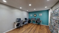 This single-room business center is thoughtfully designed to serve as a functional and visually engaging workspace. The room features rich, warm hardwood flooring that runs throughout, creating a welcoming and polished aesthetic. The walls are finished in a neutral gray, with one bold teal accent wall that adds a vibrant focal point to the space. Recessed lighting in a textured ceiling provides consistent, even illumination across the entire room.

Multiple granite-topped workstations are arranged along the walls in an L-shaped configuration, each equipped with desktop computers and ergonomic rolling chairs, accommodating several users simultaneously. A modular storage unit and a classic wall clock adorn the teal accent wall, adding both practicality and character.

A standout architectural detail is the large circular window cut through an interior wall, offering a view into an adjacent space and introducing a unique, modern design element. A French door with glass panels provides access to the connecting area. One wall is lined floor-to-ceiling with a large media and book display shelf, stocked with an extensive collection and flanked by a window with curtains that allows for natural light. A single accent chair completes the seating options, making this a versatile and well-equipped room for productivity.